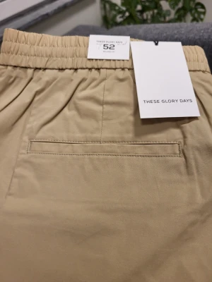 Beige shorts från These Glory Days - Snygga beige shorts från These Glory Days med elastisk midja och klassisk passform. Tillverkade i mjuk bomull, har fickor fram och en diskret bakficka. Perfekta för varma dagar och enkel att matcha med olika stilar.