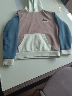 Quiksilver hoodie i pastellfärger - Snygg hoodie från Quiksilver i pastellfärger med ljusgrön huva och magficka, grå kropp och blå ärmar. Klassisk känguruficka framtill och logga på bröstet. Perfekt för en avslappnad streetwear-look.