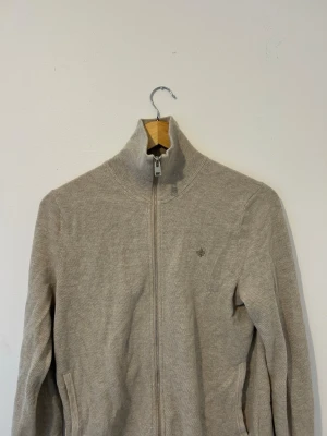 Beige stickad ziptröja med hög krage - Säljer en beige stickad tröja med dragkedja och hög krage. Tröjan har diskret broderad detalj på bröstet och två fickor framtill. Perfekt för lager på lager och enkel att matcha med olika outfits.