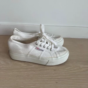 Vita Superga sneakers med platåsula - Säljer ett par klassiska vita Superga sneakers med platåsula. Skorna har en låg silhuett, vita skosnören och diskret Superga-logga på sidan. Tillverkade i canvas med gummisula för en clean och tidlös look.
