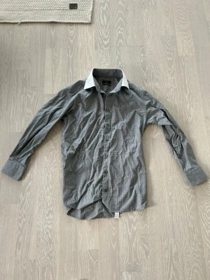 Grå rutig skjorta från Eton - Säljer en grå rutig skjorta från Eton med vit kontrastkrage och diskreta röda sömmar. Skjortan har lång ärm, knappar framtill och klassisk passform. Perfekt för dig som gillar stilrena detaljer och tidlös design. Bara att höra av sig vid minsta lilla fundering