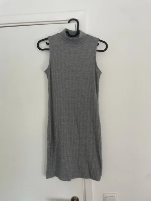 Ärmlös grå ribbad klänning - Säljer en stilren, ärmlös klänning i grått ribbat material. Klänningen har en hög krage och rak passform som ger en modern look. Perfekt för dig som gillar minimalistisk stil och vill ha något enkelt men snyggt.