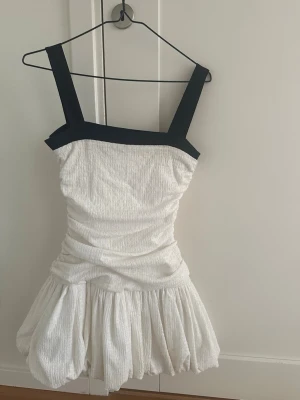 Vit och svart klänning från Zara - Supersnygg vit klänning från Zara med breda svarta axelband och kant upptill. Klänningen har en puffig kjol och draperad midja som ger en cool siluett. Materialet är strukturerat och känns lyxigt. Perfekt för dig som vill sticka ut!
