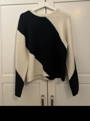 Stickad tröja med svart och vit design - Säljer en stickad tröja med unik diagonal färgblock-design i svart och vitt. Tröjan har rund halsringning och långa ärmar. Perfekt för dig som gillar en modern och stilren look.
