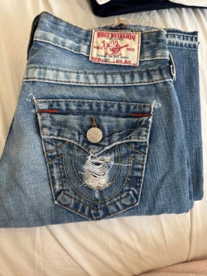 Vintage Lowwaist bootcut True Religion jeans  - Vintage bootcut True Religion jeans! Köpta i London, väldigt lowwaist. Hålet på knät är igensytt lite för det var väldigt stort. Jättesnygga detaljer på jeansen❌Står storlek W26 sitter definitivt som W25!!! ❌ Midjemått 37cm rakt över, innerbenslängd 84cm. Säljer fler snygga jeans, kolla in!