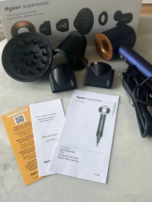 Dyson Supersonic hårfön med munstycken - Dyson Supersonic hårfön i metallicblå och guld med flera utbytbara munstycken och diffuser. Levereras med originalkartong, manualer och sladd. Modern design och avancerad teknik för snabb och skonsam hårtorkning.
