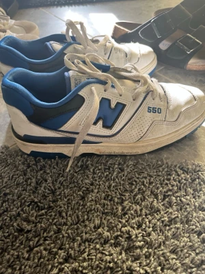 New Balance 550 vita/blå sneakers - Säljer ett par New Balance 550 sneakers i vitt och blått med svarta detaljer. Skorna har en låg silhuett, klassisk snörning och en stor N-logga på sidan. Ovandelen är i läder och mesh för en sportig vibe. Perfekta för dig som gillar retrostil.