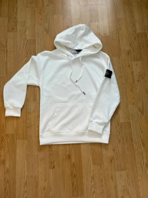 Vit hoodie från Stone Island - Säljer en clean vit hoodie från Stone Island med klassisk patch på vänster ärm. Hoodien har huva med dragsko, magficka och ribbade muddar. Perfekt för dig som gillar streetwear och vill ha en stilren look.Storlek S men passar en som är typ 168 och typ 60kg