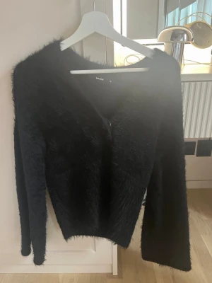 Svart fluffig kofta från Bik Bok - Mysig svart kofta från Bik Bok i fluffigt material. Koftan har långa ärmar, v-ringning och dragkedja framtill. Perfekt för dig som gillar en trendig och bekväm look med lite extra attityd.