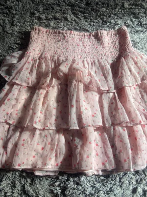 Rosa volangkjol med blommigt mönster - Supersöt kortkjol i ljusrosa med små blommor och flera volanger. Kjolen har bred smock i midjan för skön passform och är gjord i ett lätt, luftigt material som passar perfekt till vår och sommar. Perfekt för dig som gillar romantisk och feminin stil. Storlek 164 men är längre än så.