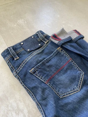 Mörkblå jeans med röda detaljer - Snygga mörkblå jeans med kontrastsömmar och röda detaljer på bakfickan. Klassisk femficksmodell med normal passform och uppvikta benslut som visar grått tyg med röda ränder. Perfekta för en avslappnad och trendig stil. Mått 📏 (cm) Totallängd: 108 Midjelängd: 38 Innersömmlängd: 82 Lårviddlängd: 27 Skador 🔧: Inga  Storlek 🧍: 31  Passform: Leonardo (Slim)  Jag kan förhandla mig till lägre pris.🙋💵 Om du har några frågor, vänligen skriv till mig 🤗