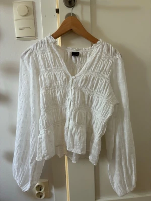 Vit blus från Gina Tricot  - Säljer en luftig vit blus med smockdetaljer och volanger. Blusen har v-ringning, knappar framtill och långa ärmar med lätt puff. I storlek S 
