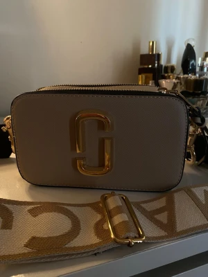 Marc Jacobs axelrämsväska - Snygg beige handväska från Marc Jacobs med guldiga metalldetaljer och ett stort logotypemblem framtill. Väskan har en bred, justerbar axelrem med text och dragkedja upptill. Perfekt för dig som vill ha en trendig och praktisk accessoar.