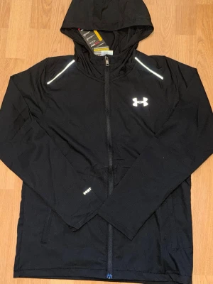 Svart vindjacka från Under Armour  -  Passar S igentligenSnygg svart vindjacka från Under Armour med huva och hel dragkedja. Jackan har reflekterande detaljer på axlarna och logga på bröstet. Tillverkad i lätt och funktionellt material, perfekt för blåsiga dagar. Två sidofickor med dragkedja.
