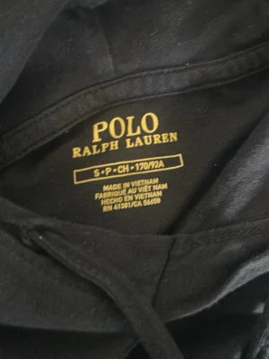 Svart hoodie från Polo Ralph Lauren - Säljer en svart hoodie från Polo Ralph Lauren med den klassiska röda broderade loggan på bröstet. Tröjan har huva och är tillverkad i mjukt material som känns skönt mot huden. Perfekt för en avslappnad och stilren look. Små märken i luvan dock.