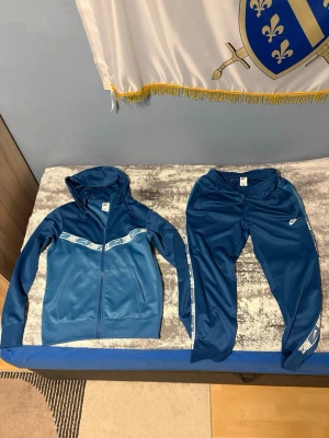 Blå Nike träningsset med huva - Säljer ett blått träningsset från Nike med zip hoodie och matchande byxor. Setet har vita Nike-loggor längs bröstet och byxsidorna, samt fickor på både jackan och byxorna. Materialet är mjukt och stretchigt, perfekt för träning eller chill.