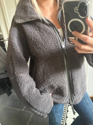 Mörkgrå teddyjacka med dragkedja - Mysig mörkgrå teddyjacka med hög krage och hel dragkedja framtill. Jackan har två sidofickor och detaljer i matchande tyg längs ärmarna. Perfekt för kyliga dagar och ger en avslappnad, trendig look. Materialet är fluffigt och mjukt, vilket gör den superbekväm.