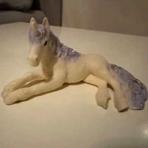 Schleich enhörning  - Säljer en söt enhörningsfigur i vit plast med lila man och svans. Figuren har liggande position och glittriga detaljer i manen. Perfekt som dekoration eller leksak för den som gillar magiska djur.
