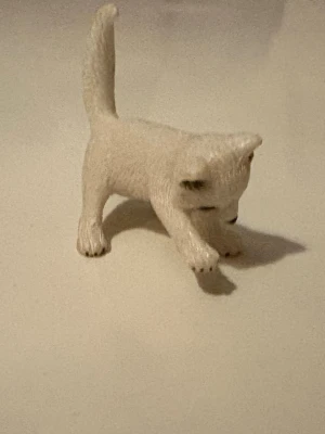 Schleich katt - Söt liten kattfigur i vit plast med detaljer som svarta nosen, öron och tassar. Figuren har en lekfull pose med ena tassen framåt och passar perfekt som dekoration eller leksak. Realistisk textur och gulligt uttryck.