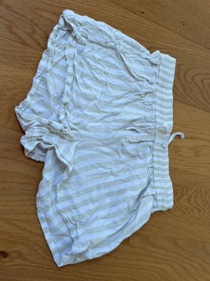 Vita randiga shorts från Serenity XS - Luftiga vita shorts från Serenity i storlek XS med tunna beigea ränder. Shortsen har bred resår i midjan och en liten snörning framtill. Tillverkade i mjukt bomullsmaterial och har en avslappnad passform med volangkant längs benen.