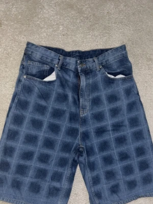 Blå rutiga jeansshorts - Säljer ett par blå jeansshorts med rutigt mönster och klassisk femficksdesign. Shortsen har bälteshällor och knappgylf. Perfekta för dig som vill sticka ut med en unik denimstil.