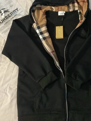 Svart hoodie med Burberry-mönster - Snygg svart hoodie från Burberry med dragkedja och klassiskt beige, svart och rött rutmönster på insidan av huvan och längs öppningen. Hoodien har fickor framtill och ribbade muddar vid ärmar och nederkant. Perfekt för en trendig och avslappnad stil.