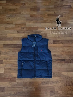 Mörkblå dunväst från Ralph Lauren - Säljer en helt ny Polo Ralph Lauren dunväst i storlek M, oanvänd med originaltaggen kvar. Klassisk marinblå vadderad modell med broderad röd Polo Pony-logga på bröstet. Hög krage, full dragkedja samt två sidofickor och en bröstficka med dragkedja. Stilren, varm och lättmatchad – perfekt över tröja eller hoodie under vår och höst. Inga fläckar, hål eller andra defekter. 📦 Snabb leverans – skickas samma dag (om ej hinder).