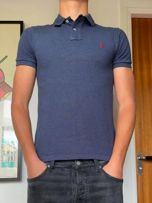 Marinblå pikétröja från Ralph Lauren - Säljer en marinblå pikétröja från Ralph Lauren med slim fit passform. Tröjan har korta ärmar, klassisk krage med knappar och den ikoniska röda loggan broderad på bröstet. Perfekt till jeans eller shorts för en clean och stilren look.