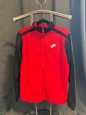 Nike ziptröja - Snygg röd ziptröja från Nike med svarta ärmar och axelpartier. Tröjan har hel dragkedja, ståkrage och två fickor framtill. Tillverkad i mjukt och bekvämt material, perfekt för träning eller chill. Ikonisk Nike-logga på bröstet.