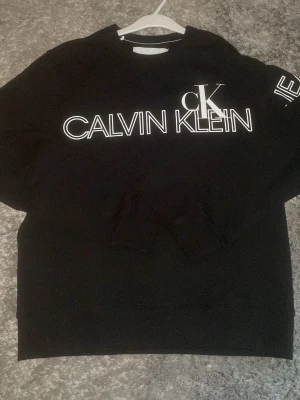 Svart Calvin Klein hoodie XS - Säljer en svart hoodie från Calvin Klein Jeans i storlek XS. Hoodien har ett stort vitt logotryck på bröstet och är tillverkad i mjukt material. Klassisk passform med ribbade muddar och rund hals. Perfekt för dig som gillar stilrena plagg med tydlig branding.