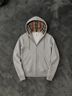 Grå hoodie från Burberry med dragkedja - Snygg grå hoodie från Burberry i storlek M. Hoodien har dragkedja framtill, kängurufickor och den klassiska Burberry-rutiga huvan. Tillverkad i mjukt material som känns skönt mot huden. Perfekt för dig som vill ha en stilren och exklusiv look.