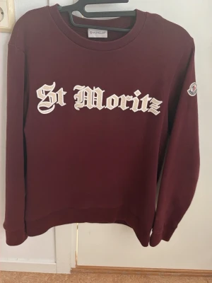 Moncler tröja - Vinröd sweatshirt från Moncler med broderad text 'St Moritz' i vitt på bröstet och Moncler-logga på ärmen. Klassisk rund halsringning och ribbade muddar. Perfekt för dig som vill ha en snygg och exklusiv tröja med streetwear-vibe.