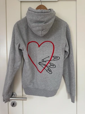 Grå hoodie med hjärta och säkerhetsnålar - Säljer en grå hoodie med huva och dragsnören. På ryggen finns ett stort rött hjärta och tre svarta säkerhetsnålar som tryck. Hoodien har en klassisk känguruficka framtill och ribbade muddar vid ärmar och midja. Perfekt för en chill och avslappnad stil.
