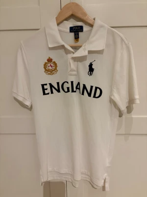 Vit pikétröja Polo Ralph Lauren England - Vit pikétröja från Polo Ralph Lauren med England-tryck och broderad logga på bröstet. Klassisk krage, korta ärmar och nummer 26 på ryggen. Använd 1 gång och köpt på kidsbrandstore. CL i juniorstorlek 