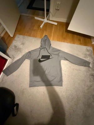 Grå hoodie med svart Nike-logga - Snygg grå hoodie från Nike med stor svart logga på bröstet och justerbar huva med svarta snören. Klassisk passform och mjukt material som känns skönt mot huden. Perfekt för en avslappnad och sportig stil.