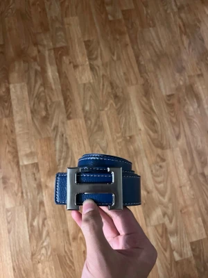 Blått skärp med silverfärgat H-spänne - Snyggt blått bälte med ett stort, silverfärgat H-format spänne. Bältet har vita kontrastsömmar längs kanterna och är gjort i ett slätt material som liknar skinn. Perfekt för att lyfta vilken outfit som helst.