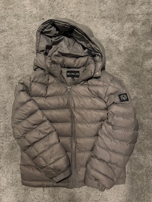Grå dunjacka med huva från Evaldi - Säljer en grå pufferjacka från Bvaldi med stor huva och dragkedja framtill. Jackan har quiltad design, två fickor med dragkedja och en patch på ärmen. Perfekt för kalla dagar och riktigt snygg till alla sorts stilar. Köpt för 2999kr på bvaldis egna hemsida. Skicket är 9,5 av 10 använd max 3 gånger då jag beställde en storlek för liten och den blev för liten direkt. Kom gärna med bud eller frågor