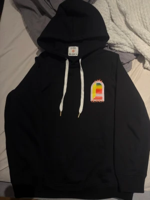 Svart hoodie från Casablanca - Svart hoodie från Casablanca med stor färgglad grafik på ryggen och mindre tryck på bröstet. Huva med vita snören och avslappnad passform. Perfekt för dig som gillar streetwear och vill sticka ut med cool design.