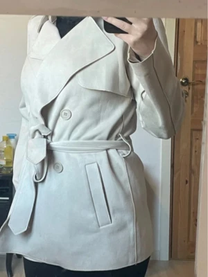 Ljusbeige kort trenchcoat med skärp - Snygg ljusbeige trenchcoat med dubbelknäppning och tillhörande midjebälte. Jackan har bred krage, två framfickor och en kortare, modern passform. Perfekt för dig som vill ha en stilren och klassisk look. Materialet ser ut att vara mjukt och matt.