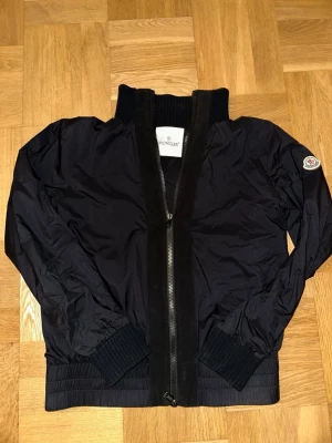 Svart vindjacka från Moncler - Snygg svart vindjacka från Moncler med hög krage och ribbade muddar. Jackan har dragkedja framtill och Moncler-logga på ärmen. Tillverkad i ett lätt, glansigt material som står emot vind. Perfekt för dig som vill ha en stilren och exklusiv look. Kan skicka fler bilder vid behov för äkthet.