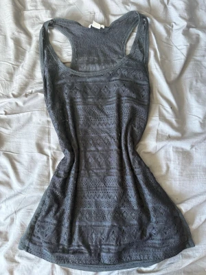 Svart spetslinne med broderade detaljer - Snyggt grått linne med broderade spetsdetaljer och mönstrad framsida. Linnet har ärmlös design och brottarrygg, vilket ger en sportig men ändå feminin look. Perfekt att matcha med jeans eller kjol för en trendig stil.