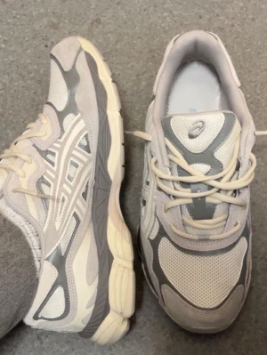Vita och grå Asics sneakers - Snygga sneakers från Asics i vitt och ljusgrått med klassisk mesh och syntetdetaljer. Skorna har en sportig vibe med tydliga Asics-loggor på sidorna och tungan. Perfekta för dig som gillar en clean och stilren look.