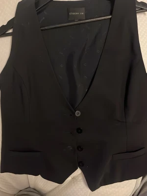 Svart kostymväst från STOCKH LM - Klassisk svart kostymväst från STOCKH LM med v-ringad hals och fyra knappar framtill. Västen har två fickor och är perfekt för att lyfta din outfit till nästa nivå. Snygg att bära över en skjorta eller t-shirt.
