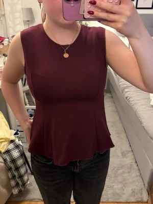 Vinröd ärmlös topp med utsvängd midja - Säljer en vinröd ärmlös topp med rund halsringning och lätt utsvängd midja. Toppen har en enkel och stilren design som passar perfekt till jeans eller kjol. Materialet känns mjukt och följsamt mot kroppen.