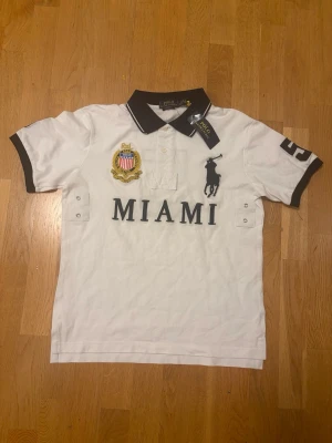 Vit piké med Miami-tryck från Polo Ralph Lauren - Vit pikétröja från Polo Ralph Lauren med svart krage och ärmslut, stort MIAMI-tryck framtill, broderad logga och nummer 5 på ärm och rygg. Klassisk polodesign med knappar vid halsen och detaljerade broderier.