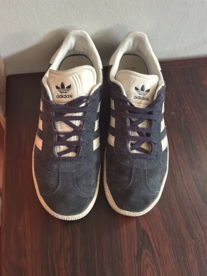 Adidas Gazelle mörkblå sneakers - Säljer ett par klassiska Adidas Gazelle sneakers i mörkblå mocka med vita detaljer och de ikoniska tre ränderna på sidorna. Skorna har vit sula och plös samt guldfärgad Gazelle-text på sidan. Perfekta för dig som gillar retrostil och streetwear. Pris kan diskuteras hör av er vid frågor.
