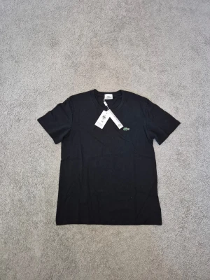 Lacoste - Klassisk svart t-shirt från Lacoste med den ikoniska gröna krokodilloggan broderad på bröstet. T-shirten har rund hals och korta ärmar, tillverkad i mjuk bomull för en skön känsla. Perfekt basplagg med clean look.