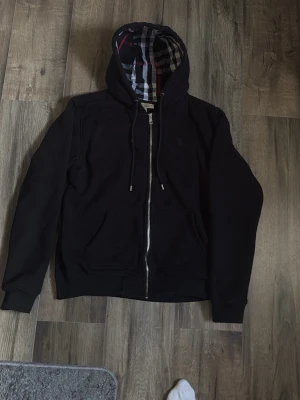 Svart hoodie med rutigt foder - (L men passar rätt så bra som M med)Snygg svart hoodie från Burberry med dragkedja framtill och klassiskt rutigt foder i huvan. Tröjan har dragsko i huvan, två fickor och diskret broderad logga på bröstet. Perfekt för dig som gillar stilrena detaljer och premiumkänsla.