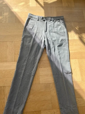 Gråa raka kostymbyxor - Snygga grå kostymbyxor med raka ben och pressveck. Byxorna har bälteshällor, sidofickor och en bakficka med knapp. Materialet ser ut att vara en blandning av bomull och polyester, vilket ger en klassisk och stilren look.                                 Storlek 30/30.                                                               Pris kan diskuteras 