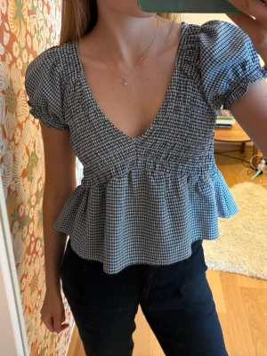 Rutig blus med puffärm och v-ringning - Blus med svartvitt rutigt mönster, puffärmar och v-ringad hals. Modellen har en volang nedtill och smockdetaljer vid ärmslut. 